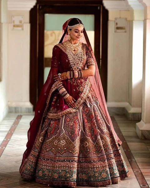 Bride wearing a deep maroon bridal lehenga with intricate embroidery, paired with traditional jewellery and dupatta, posing gracefully in a heritage-style corridor.